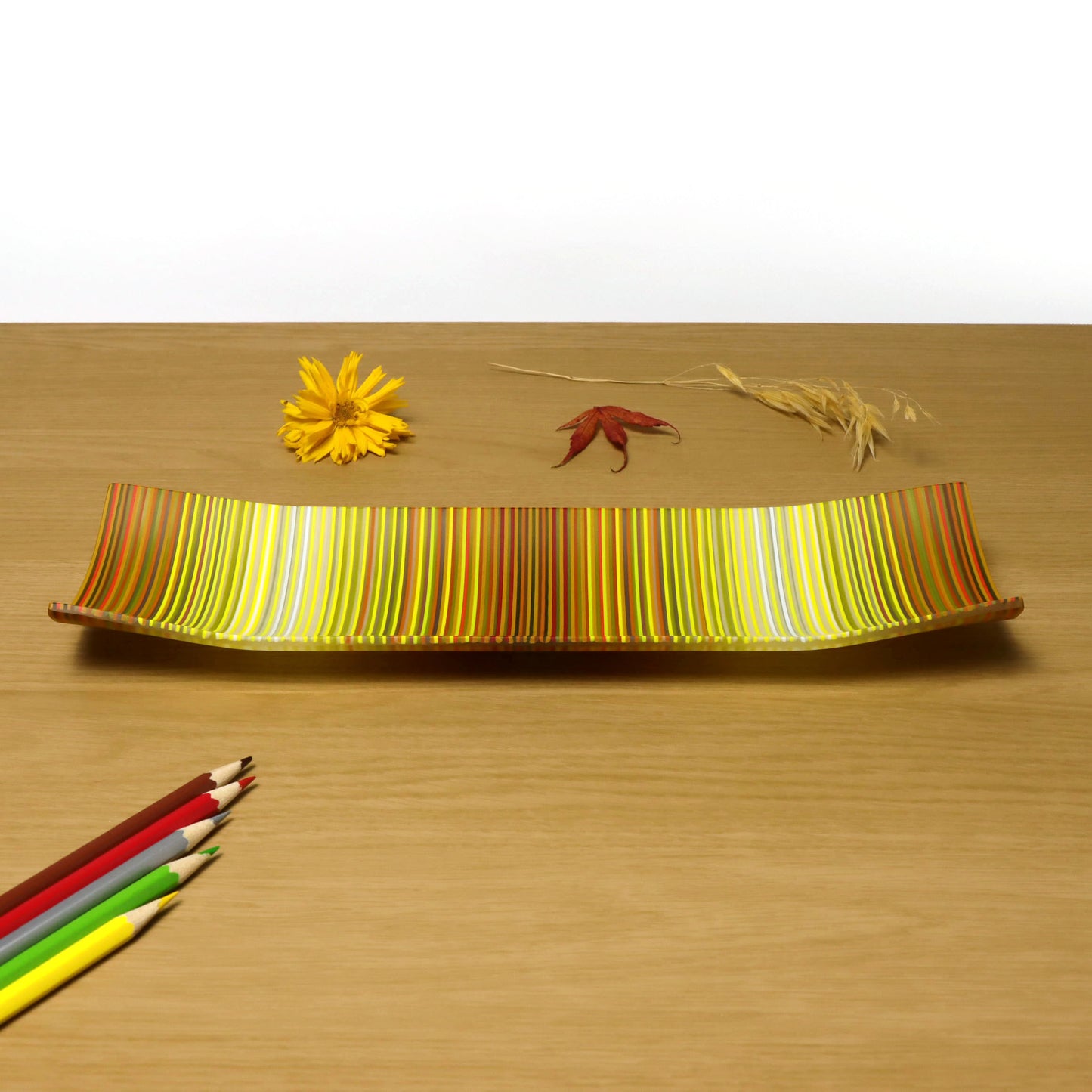 A vibrant ColourWave Glass fused glass plate with a pattern of alternating vertical stripes in yellow, orange, and red. The rectangular plate features elegantly curving edges that rise gracefully at the corners. It is presented on a wooden surface, accentuated by a yellow flower, a red leaf, and strands of wheat to the right, with an array of coloured pencils in the foreground.