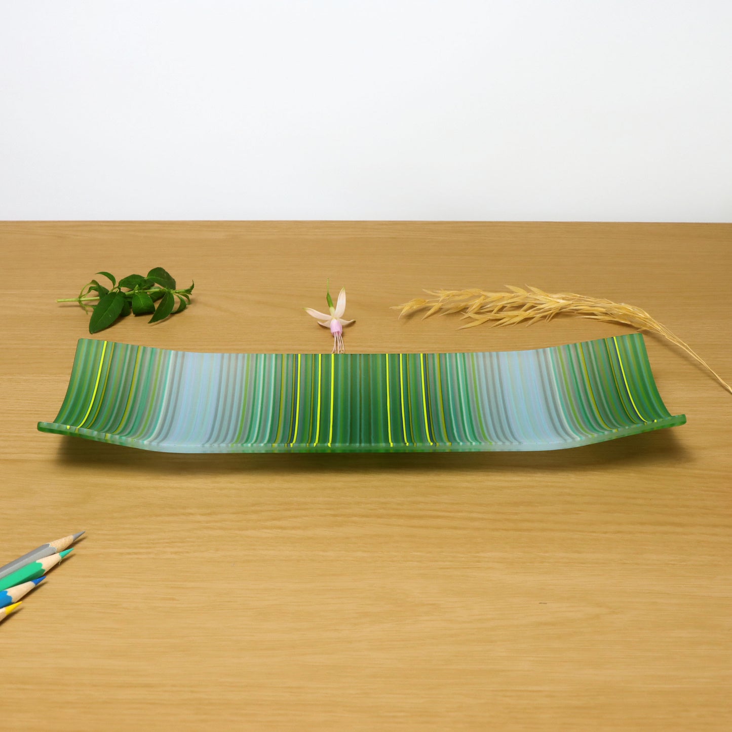 A decorative shallow rectangular boat shaped glass plate with raised corners and a colourful ColourWave stripe patterns showing key colours of green and pale blueA rectangular fused glass plate from ColourWave Glass, featuring a striking array of vertical stripes in shades of blue and green. The plate’s design includes corners that rise gently, giving it a sophisticated, three-dimensional shape.