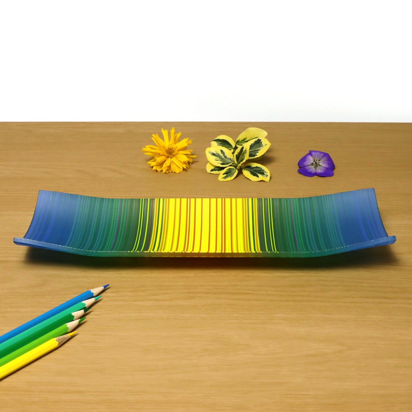 A ColourWave Glass fused glass plate, displaying a beautiful gradient that transitions from blue to yellow. The plate’s rectangular shape gracefully rises at the corners, creating an elegant raised edge. It rests on a wooden surface, complemented by a yellow flower, green leaves, and a purple flower above, with a collection of coloured pencils below, highlighting its artistic presentation.