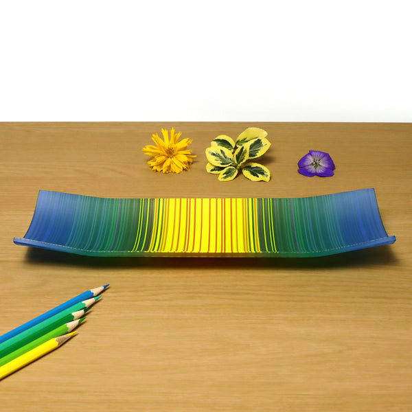 A ColourWave Glass fused glass plate, displaying a beautiful gradient that transitions from blue to yellow. The plate’s rectangular shape gracefully rises at the corners, creating an elegant raised edge. It rests on a wooden surface, complemented by a yellow flower, green leaves, and a purple flower above, with a collection of coloured pencils below, highlighting its artistic presentation.