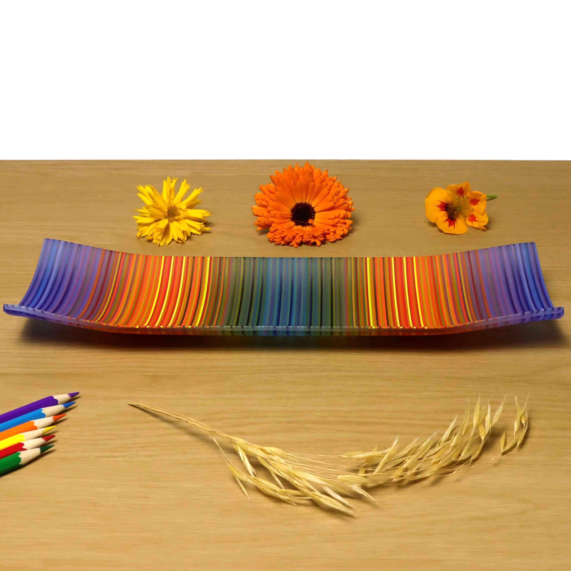 A ColourWave Glass fused glass plate, featuring a spectrum of colours including blue, orange and teal green in vertical stripes, with many more colours seen up  close. The plate has corners that elegantly curve upwards and is placed on a wooden surface, highlighted by colourful flowers and wheat stalks in the foreground, and coloured pencils placed below.