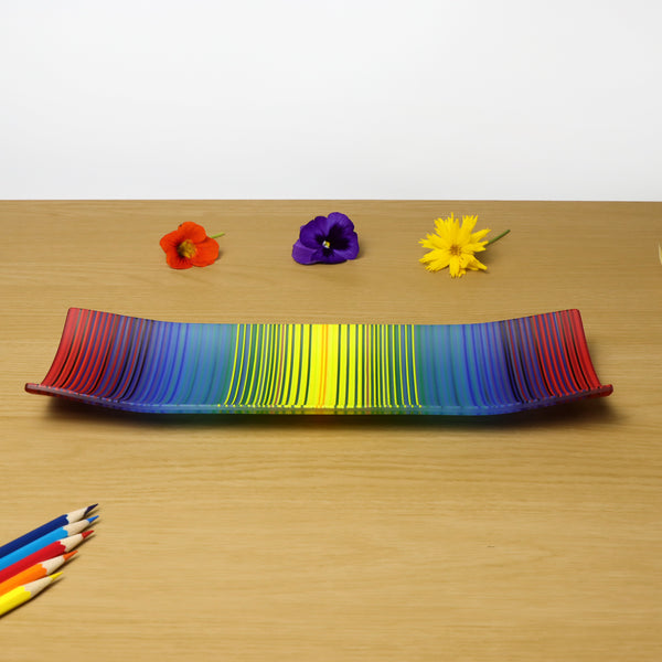 A ColourWave Glass fused glass plate, showcasing a vibrant spectrum of colours that transition smoothly from one to another, reminiscent of a rainbow just not in the same order!. The plate’s design features gracefully curving corners that add a touch of elegance to its rectangular shape. It is placed on a wooden surface, which enhances the visual appeal of the glasswork’s rich hues along with other decorative items to enhance its appeal.