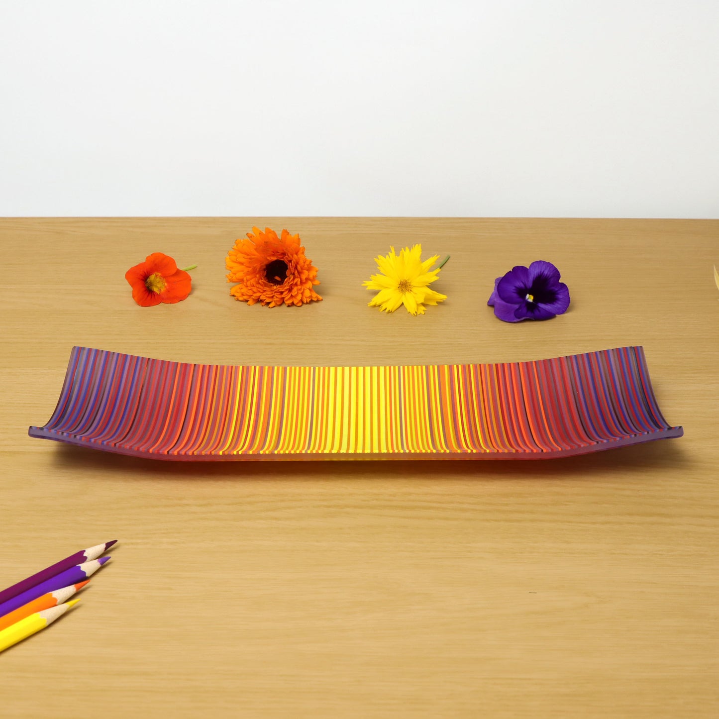 A ColourWave Glass fused glass plate, rectangular in shape, displays vibrant stripes in shades of purple, red, orange, and yellow. The plate is distinctively curved upwards at each corner, forming an elegant raised edge. It rests on a wooden surface, complemented by a colourful arrangement of flowers and a set of pencils that match the plate’s vivid stripes.