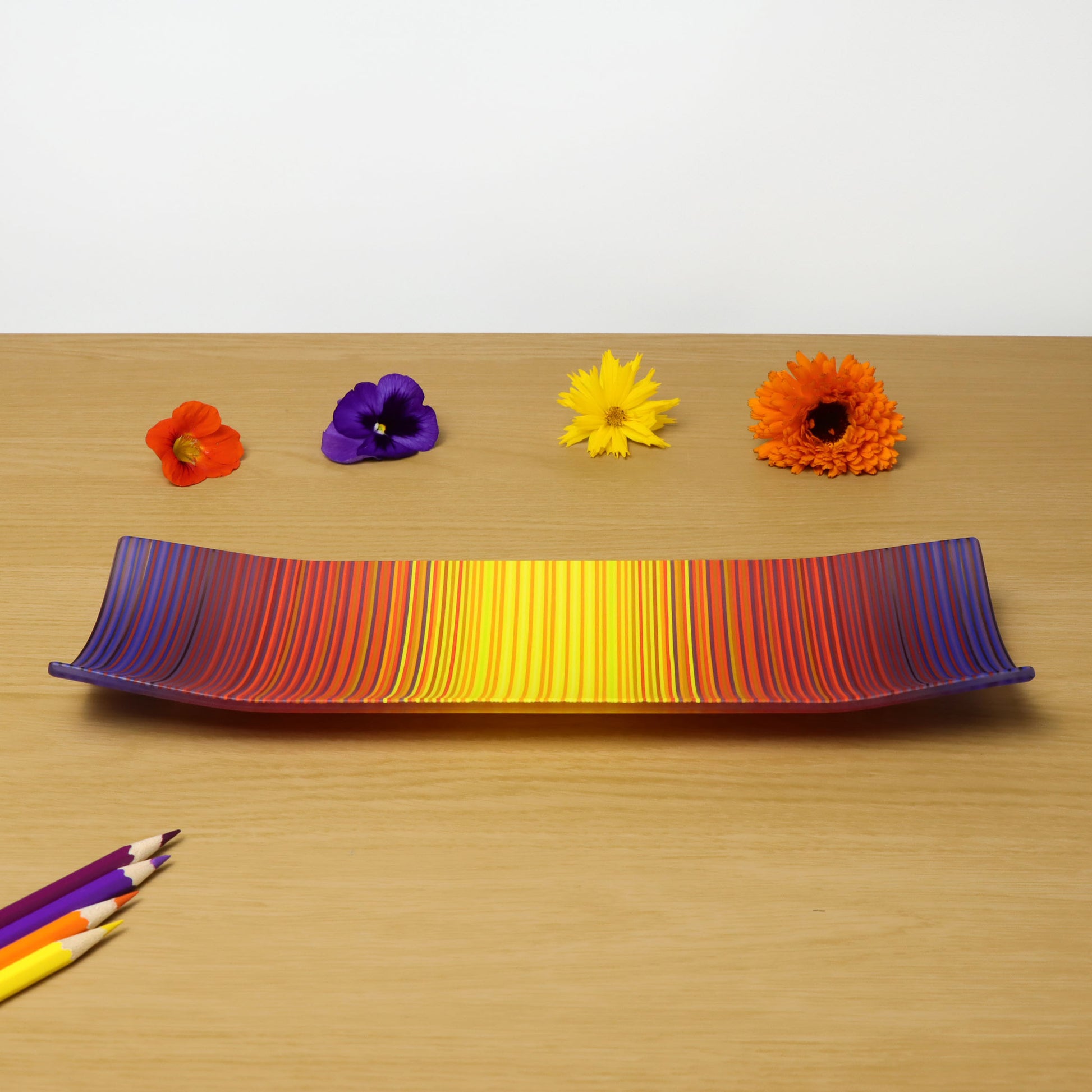 A ColourWave Glass fused glass plate, showcasing a spectrum of colours that blend from deep purples to bright yellows. The rectangular plate has a gentle curvature at the corners, enhancing its elegant and contemporary design. It is presented on a wooden surface, accompanied by colourful flowers and pencils that complement its vibrant hues.