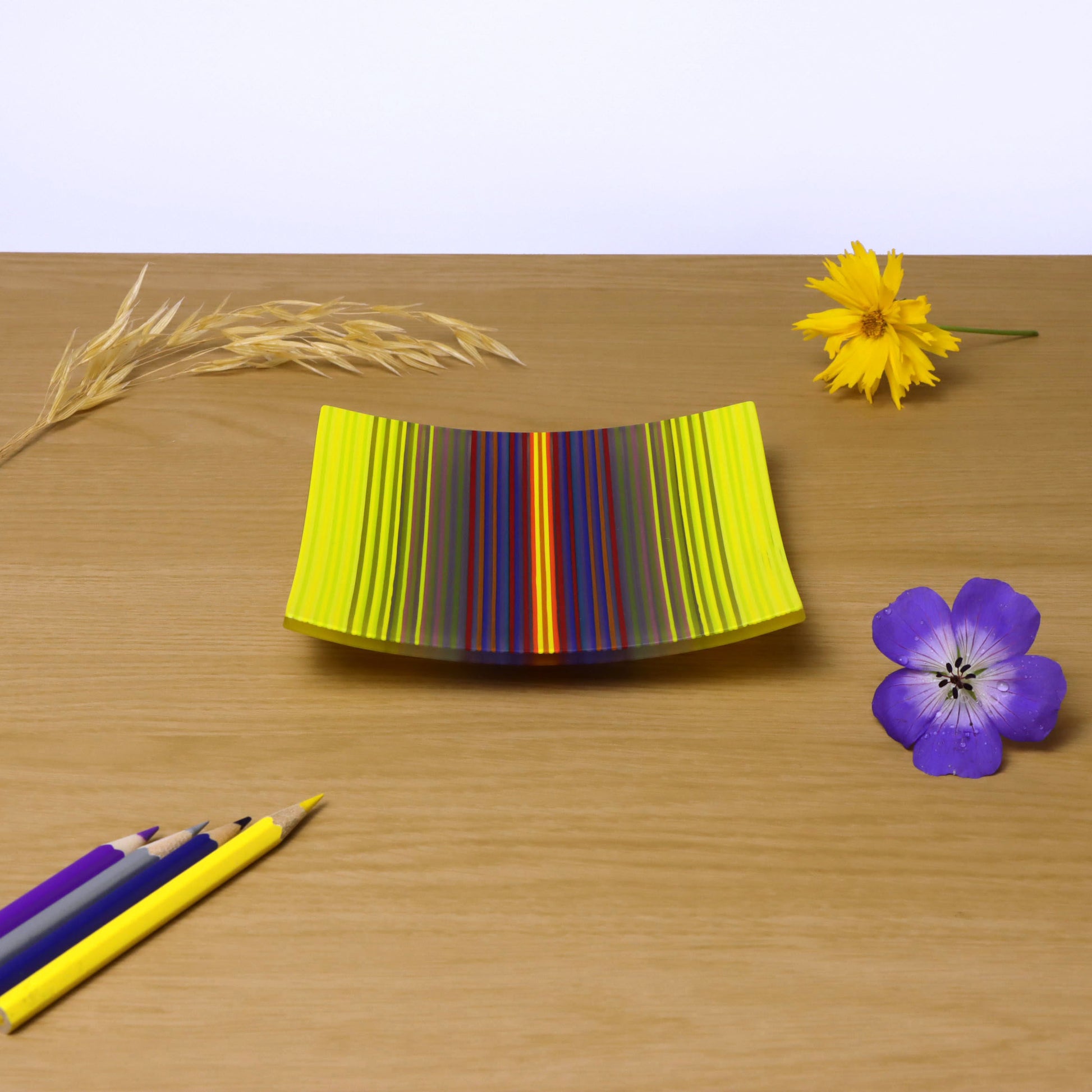 A ColourWave fused glass plate, resting on a wooden surface, displays a captivating vibrant gradation from yellow to purple, with many other colours making up the sequence. Adjacent to the plate, a yellow flower and a single stalk of wheat add a touch of nature to the scene. In the foreground, coloured pencils in matching shades of yellow purple and blue, lie diagonally pointing towards the plate, with a striking purple flower adjacent.