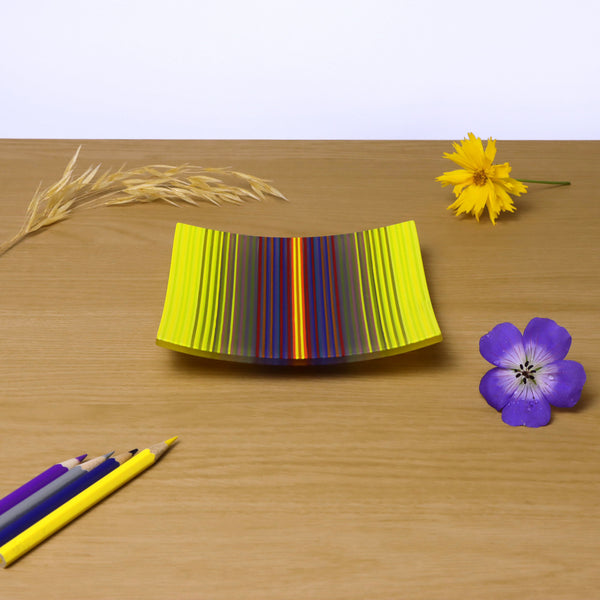 A ColourWave fused glass plate, resting on a wooden surface, displays a captivating vibrant gradation from yellow to purple, with many other colours making up the sequence. Adjacent to the plate, a yellow flower and a single stalk of wheat add a touch of nature to the scene. In the foreground, coloured pencils in matching shades of yellow purple and blue, lie diagonally pointing towards the plate, with a striking purple flower adjacent.