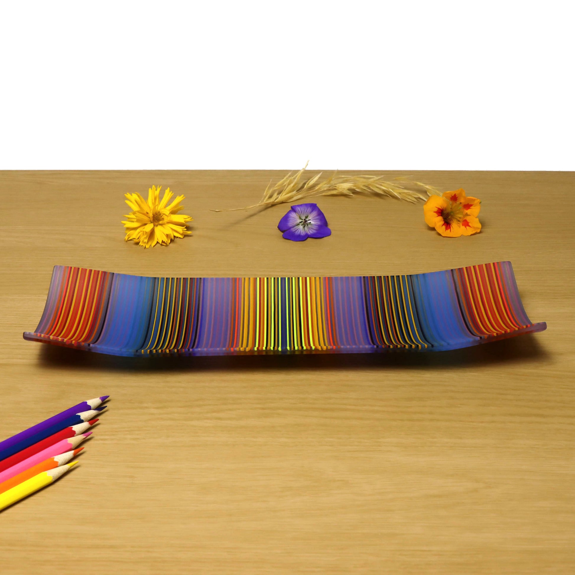 A rectangular glass plate with vibrant, vertical ColourWave stripes in a spectrum of colours including orange, yellow, green, blue, and purple. The plate features a gentle upward curve at each corner and is displayed on a wooden surface. Adjacent to the plate are colourful pencils aligned in a similar colours, and behind the plate are a few natural elements such as wheat stalks and flowers in yellow, purple, and orange.