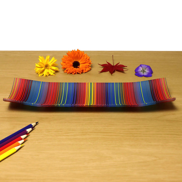 A ColourWave Glass fused glass plate, boasting a vibrant pattern of vertical stripes in a medley of colours including red, blue, yellow, and green. The rectangular plate features a subtle upward curve at each corner, lending an elegant raised edge to its design. The plate is presented on a wooden surface, complemented by a bright orange Gerbera daisy, a red maple leaf, a purple pansy flower, and an assortment of coloured pencils arranged in the foreground.