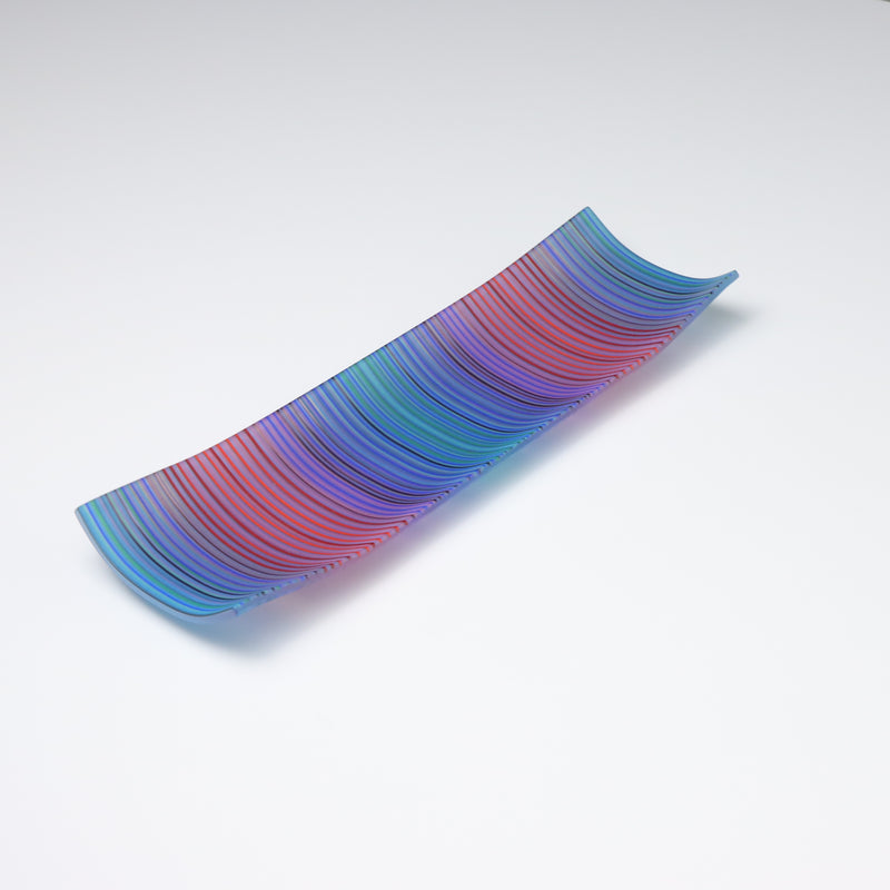 A rectangular ColourWave Glass fused glass plate displaying a vibrant array of vertical stripes in shades of blue, green, red, and purple. The plate exhibits a subtle upward curve at each corner, lending it a sophisticated and contemporary appearance. Its smooth surface catches and reflects the light, showcasing the rich colours and fine strands of glass used to create this piece. Positioned against a white backdrop, the plate’s vivid colours and stylish design are prominently featured.