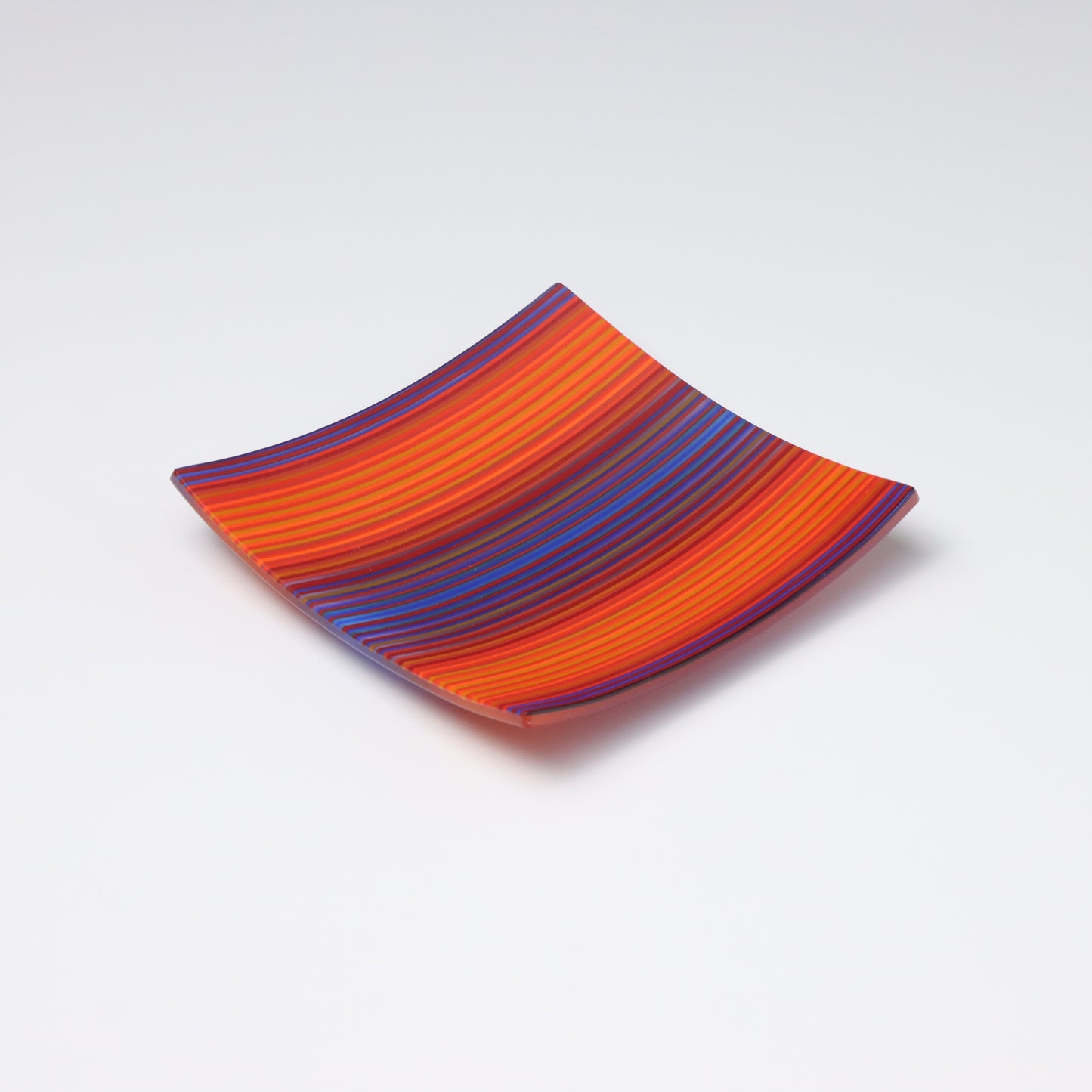 An exquisite ColourWave fused glass plate, square in shape with corners that subtly curve upwards to form a shallow dish. The plate is decorated with a lively pattern of red, orange, and blue stripes, in a wave like pattern. Set against a  white background, the plate’s vibrant colours are beautifully accentuated.