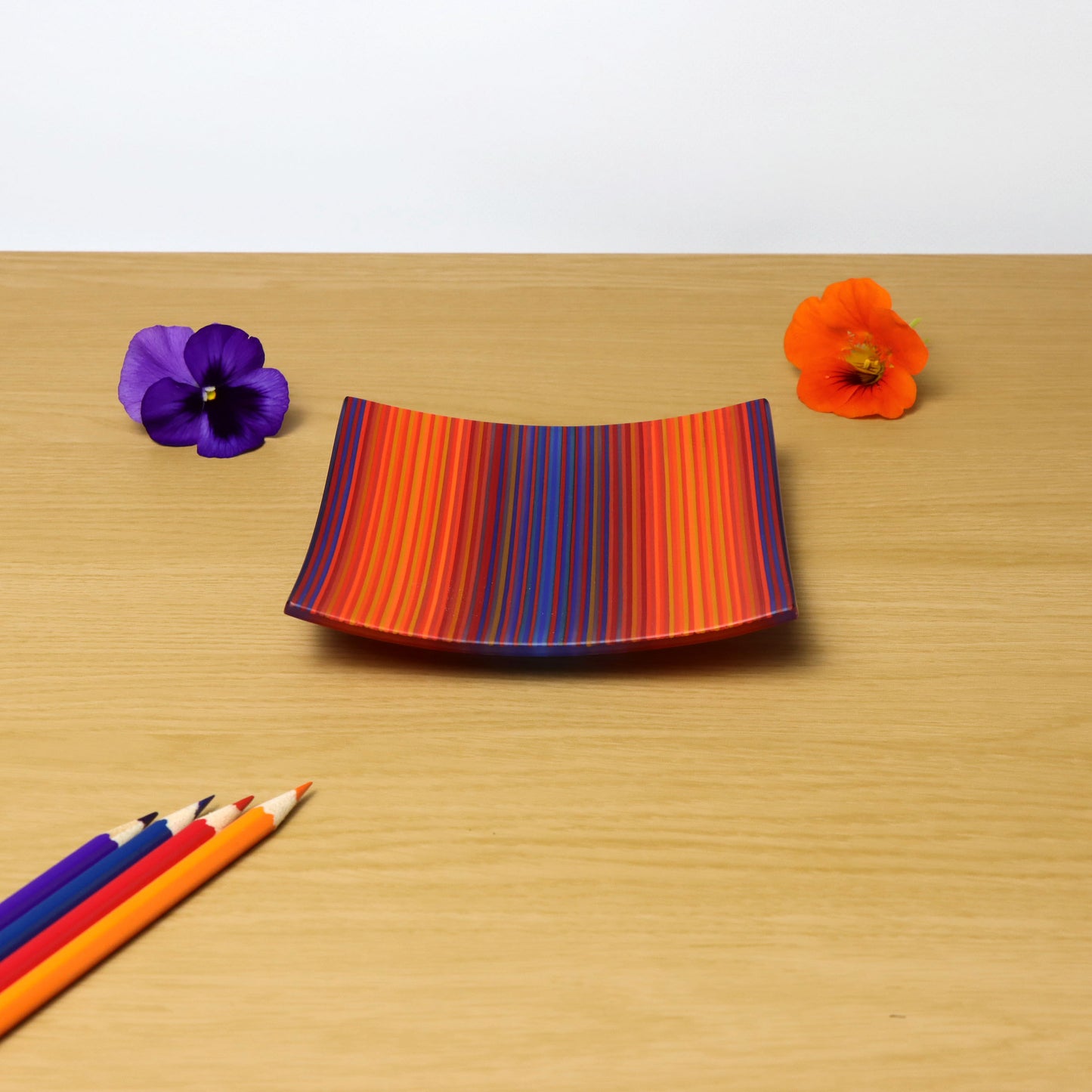 A captivating ColourWave fused glass plate, square in shape, showcasing vibrant stripes in shades of red, orange, purple, and blue. The plate is situated on a wooden surface, offering a warm contrast. To the right of the plate, two flowers—one purple, one orange—add a natural touch to the setting. In the bottom left corner, a set of coloured pencils with their tips pointing towards the plate, echoing the plate’s vivid colour scheme.