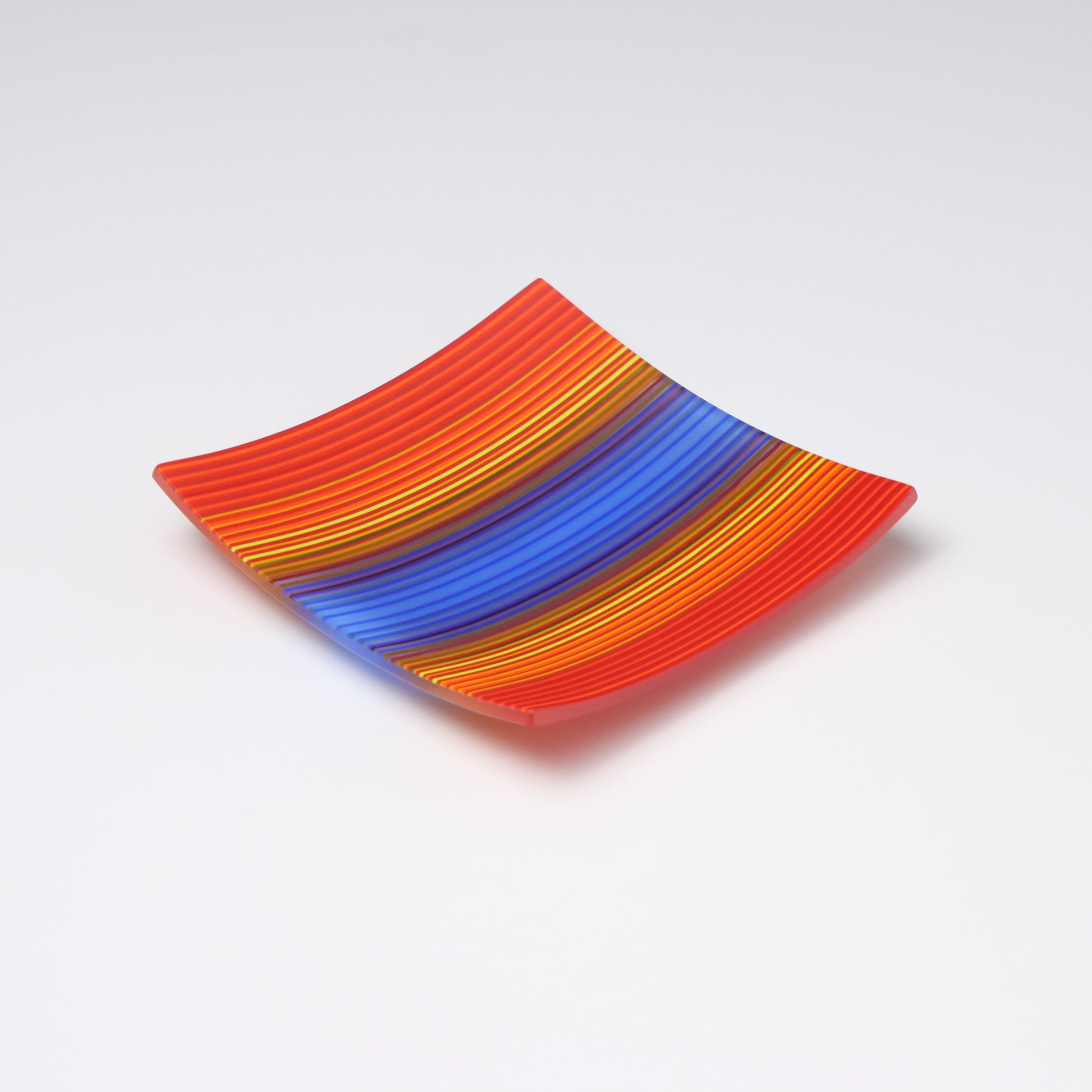 A ColourWave Glass fused  glass plate featuring a vibrant array of stripes in shades of red, orange, blue, and yellow. The plate has a square shape with upturned corners and is set against a plain white background. The pattern and the bright colours create an eye-catching visual effect that showcases the intricate craftsmanship of fused glass technique in the UK