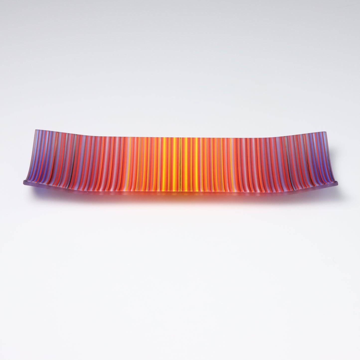 A ColourWave Glass fused glass plate displaying a striking pattern of vertical stripes in shades of red, orange, purple, and blue. The rectangular plate features corners that gently curve upwards, forming a sophisticated and contemporary shape. The plate’s smooth surface catches the light, emphasizing the intensity and beauty of the colours. It is presented against a stark white background, which serves to highlight its bold colours and refined design.