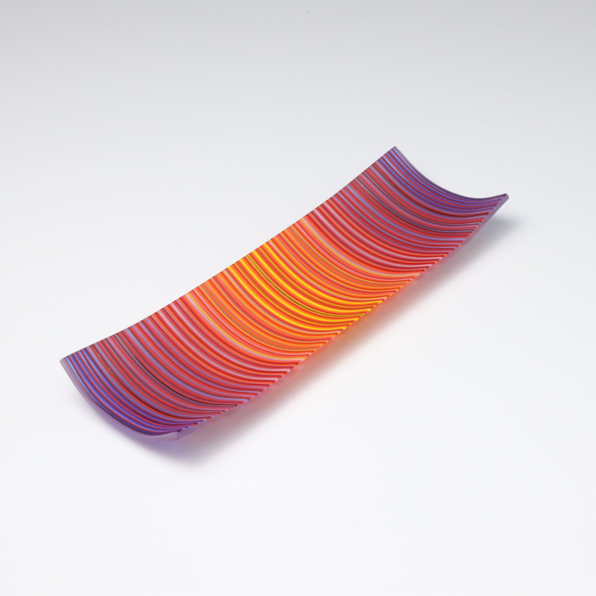 A ColourWave Glass fused glass plate displaying a striking pattern of vertical stripes in shades of red, orange, purple, and blue. The rectangular plate features corners that gently curve upwards, forming a sophisticated and contemporary shape. The plate’s smooth surface catches the light, emphasizing the intensity and beauty of the colours. It is presented against a stark white background, which serves to highlight its bold colours and refined design.