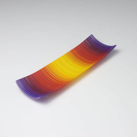 A rectangular ColourWave Glass fused glass plate showcasing a vivid spectrum of colours, with purple at the edges blending into red, orange, and yellow towards the centre. The plate has a smooth surface and its corners gently curve upwards, giving it a contemporary and elegant profile.