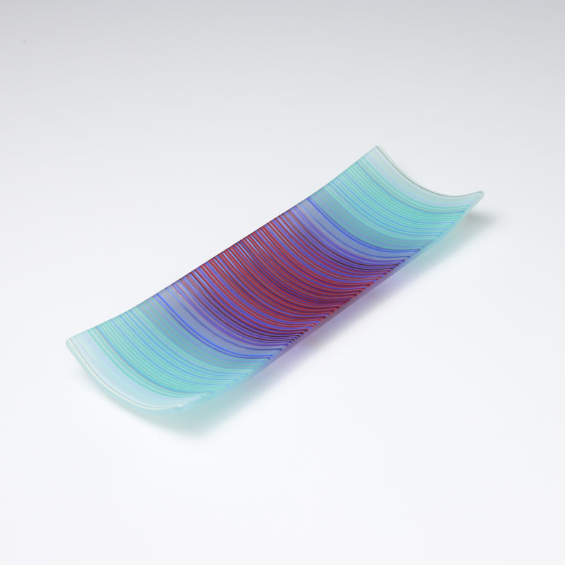 A rectangular ColourWave Glass fused glass plate, boasting a vibrant gradient from turquoise to deep blue and purple in vertical stripes. The plate’s corners elegantly curve upwards, adding a modern twist to its design. The smooth transition of colours offers a serene visual experience, while the craftsmanship reflects a high level of skill in glass work