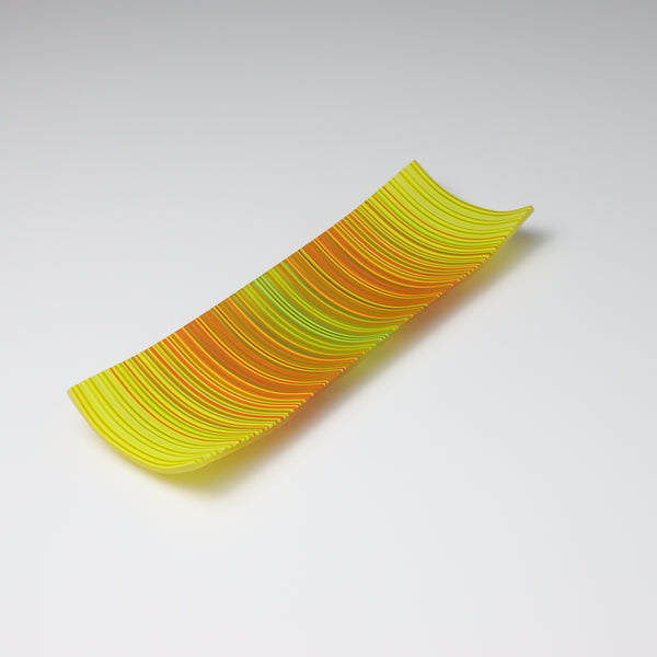 A ColourWave Glass fused glass plate, adorned with a vibrant pattern of vertical stripes in shades of yellow, green, orange, and red. The rectangular plate gently curves at the corners, forming a shallow bowl shape. The bright colours and sleek design emphasize its role as an attractive decorative piece.