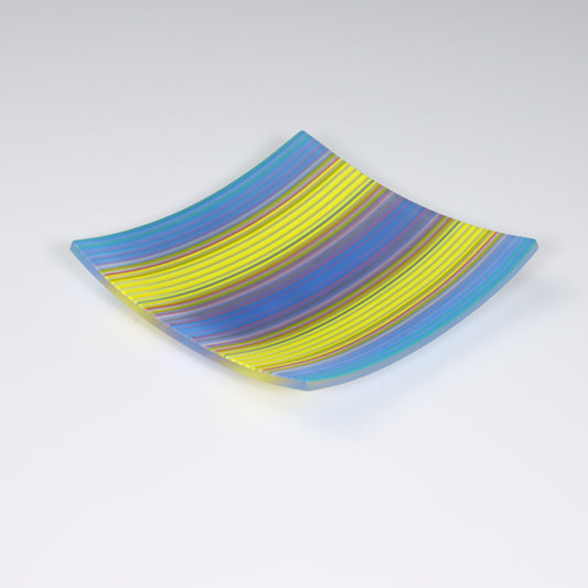 A square-shaped fused glass plate with corners that curveupwards, showcasing a ColourWave Glass design. The plate displays parallel stripes in a spectrum of colours including shades of blue, yellow, with others seen up close. The stripes curve and flow across the surface, giving the impression of movement. The glass has a lustrous finish and is set against a muted grey background, which highlights the plate’s vivid colours.