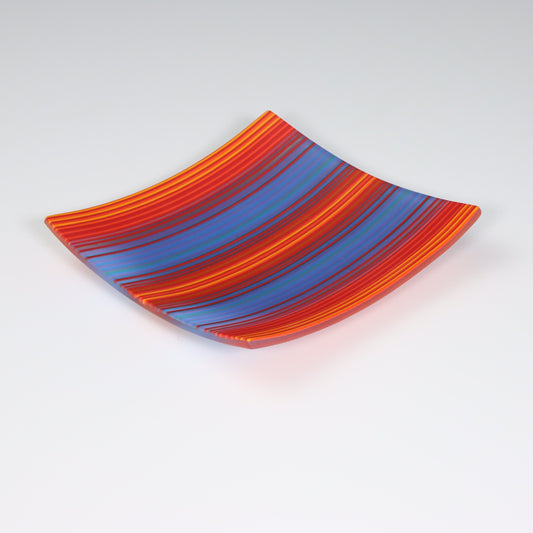 A square-shaped ColourWave fused glass plate displaying a pattern of stripes in bold red, blue, and orange hues. The plate’s corners curve upwards, forming a shallow bowl that captures the light, accentuating the vivid colours. This piece exemplifies the vibrancy of fused glass, serving as a visually striking decorative item.
