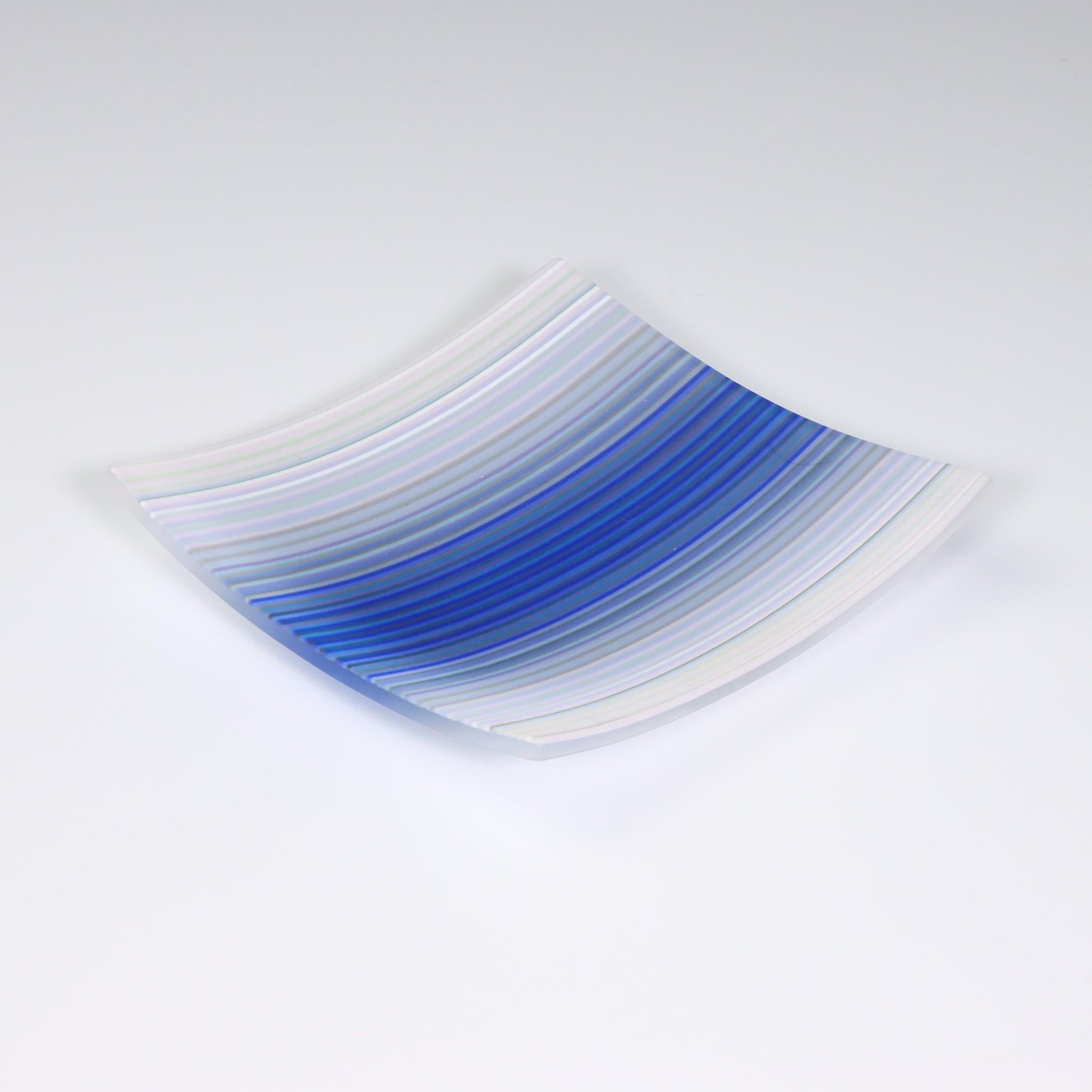 A square shaped ColourWave Glass fused glass plate, exhibiting a striking design. The colour transitions from a tranquil white to an array of blues, culminating in a deep navy in the center. The modernity of the plate’s form is complemented by the vibrant colour palette, evoking a sense of nature’s serenity. The piece is set against a contrasting light grey background, which serves to highlight its vivid colours.