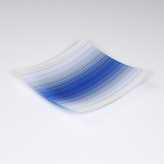 A square shaped ColourWave Glass fused glass plate, exhibiting a striking design. The colour transitions from a tranquil white to an array of blues, culminating in a deep navy in the center. The modernity of the plate’s form is complemented by the vibrant colour palette, evoking a sense of nature’s serenity. The piece is set against a contrasting light grey background, which serves to highlight its vivid colours.