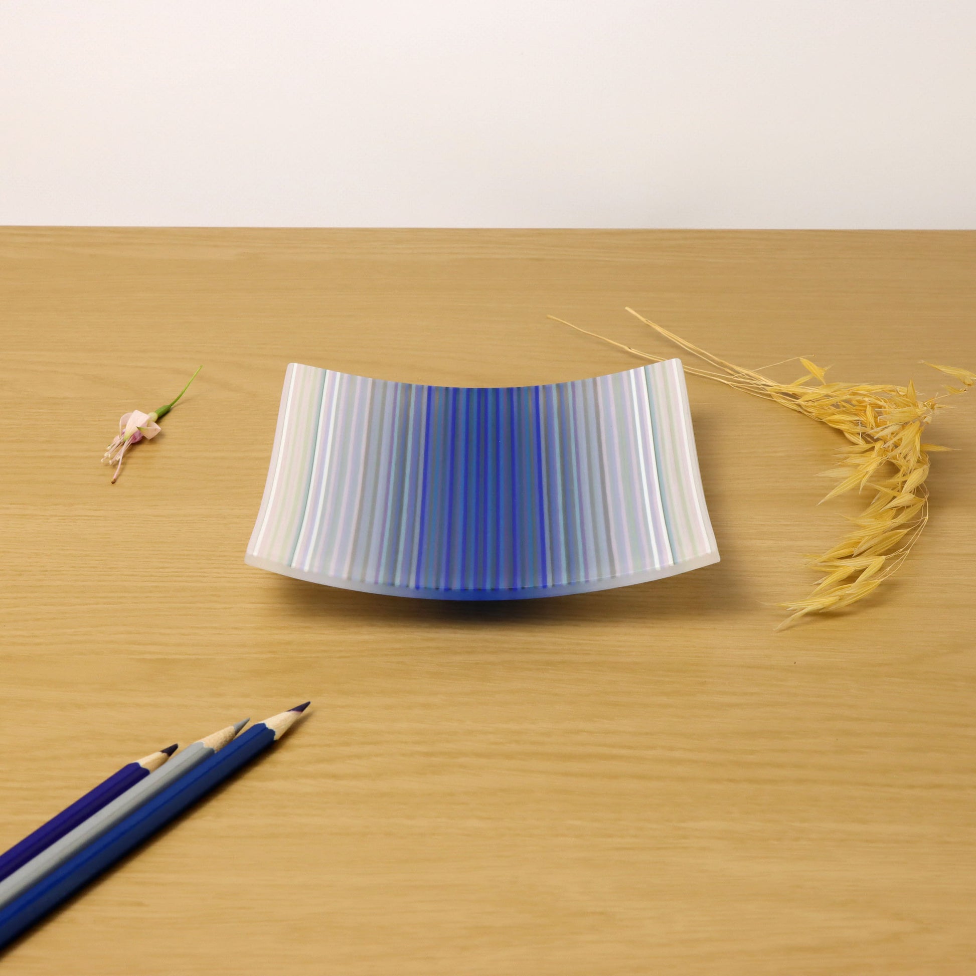 A ColourWave Glass fused glass plate, beautifully displayed on a wooden surface. The plate is adorned with vertical stripes in a spectrum of blues, forming a wave like pattern. The plate’s gentle curvature adds depth to its form, enhancing the visual impact of the ColourWave design. Coloured pencils in toning colours, placed in front of the plate, harmonise with the colour scheme of this fused glass piece.