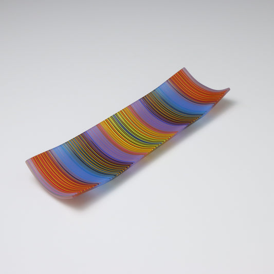 An exquisite ColourWave Glass plate with a rectangular shape, featuring a striking pattern of vertical stripes in a bold colour palette including shades of blue, purple, orange, yellow and green, with many more seen up close. The design is complemented by the plate’s corners, which gracefully curve upwards, giving it a refined and elevated appearance.