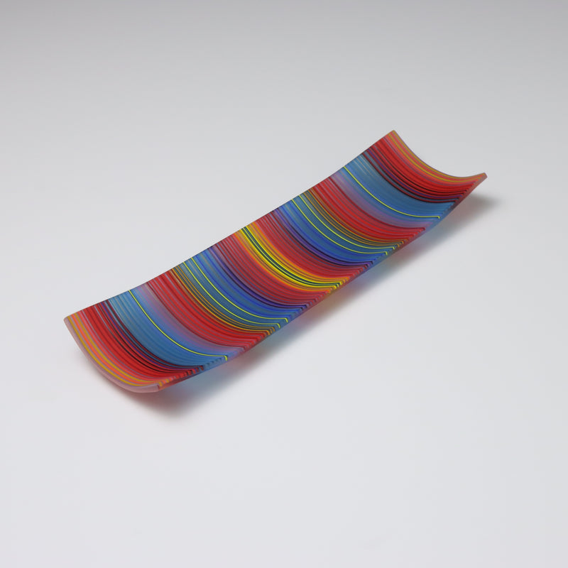 A ColourWave Glass fused glass plate, arrayed with vertical stripes in vibrant red, blue, yellow, and green. The plate’s rectangular shape is enhanced by the corners that curve upward, forming a shallow, dish-like appearance. Set against a grey background, the plate’s rich stripes and distinctive design are highlighted, making it an eye-catching piece.