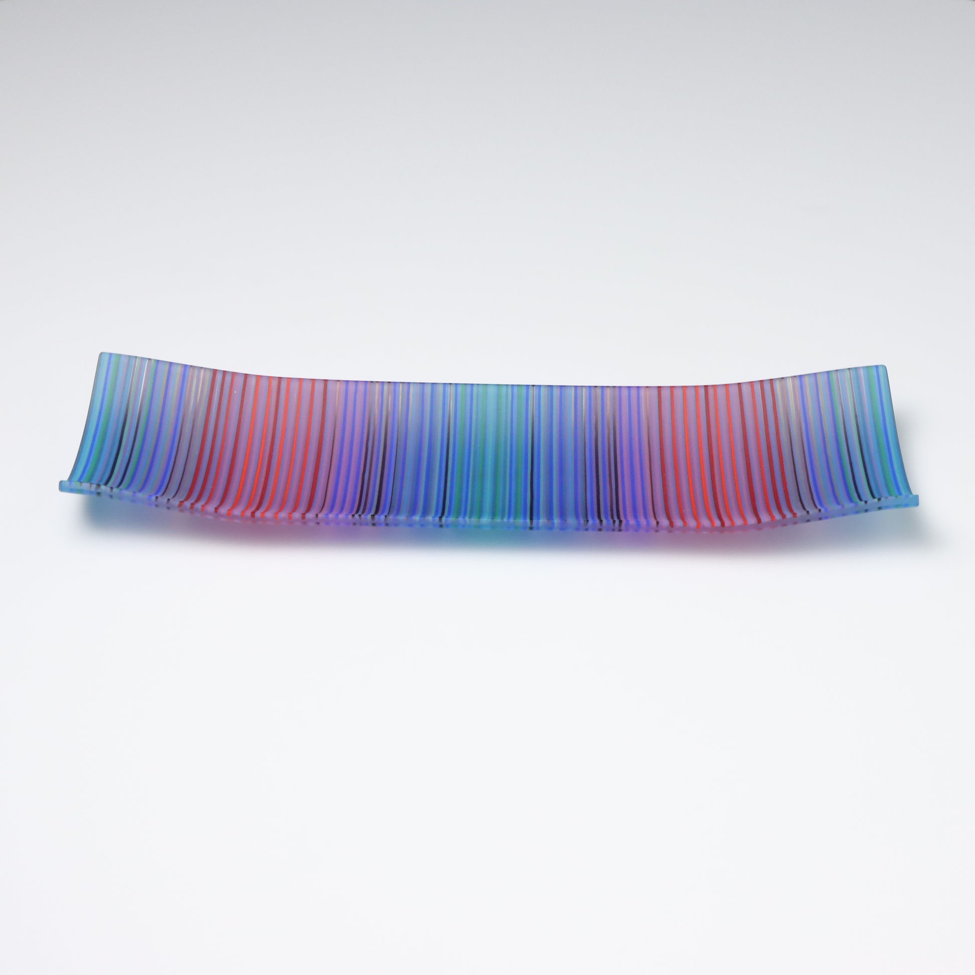 A rectangular ColourWave Glass fused glass plate displaying a vibrant array of vertical stripes in shades of blue, green, red, and purple. The plate exhibits a subtle upward curve at each corner, lending it a sophisticated and contemporary appearance. Its smooth surface catches and reflects the light, showcasing the rich colours and fine strands of glass used to create this piece. Positioned against a white backdrop, the plate’s vivid colours and stylish design are prominently featured.
