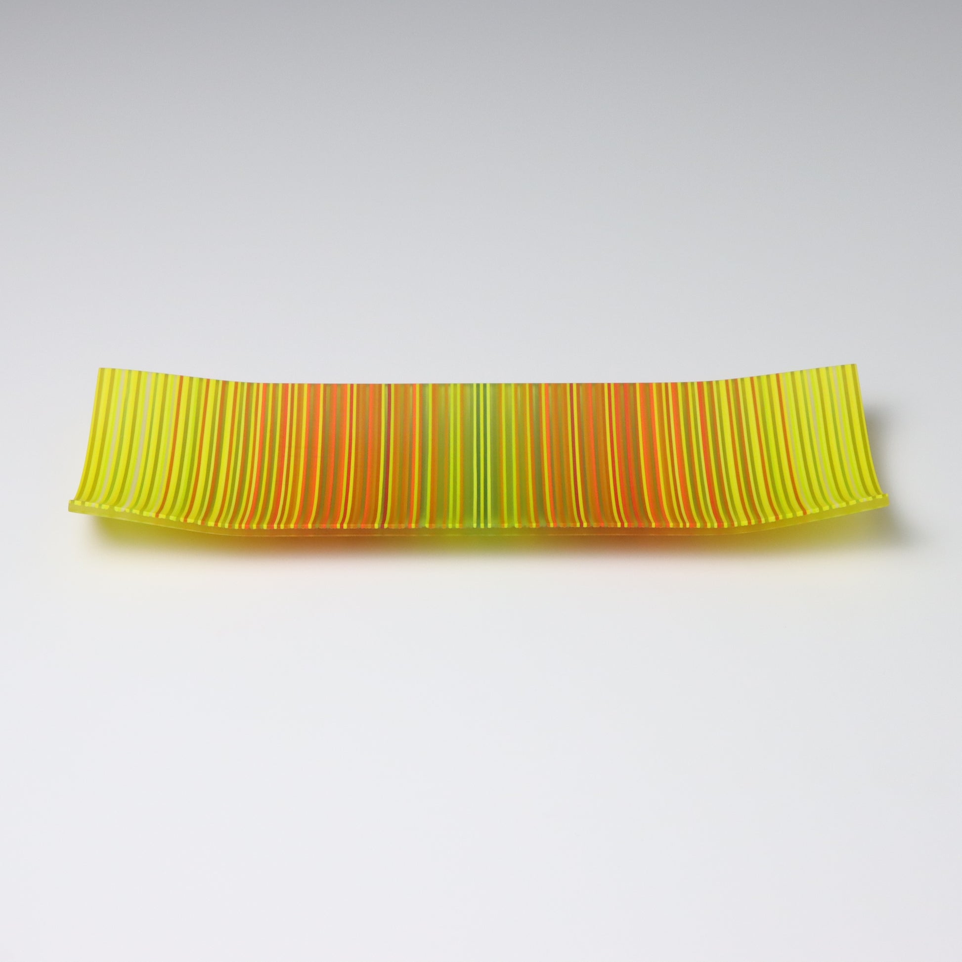 A ColourWave Glass fused glass plate, adorned with a vibrant pattern of vertical stripes in shades of yellow, green, orange, and red. The rectangular plate gently curves at the corners, forming a shallow bowl shape. The bright colours and sleek design emphasize its role as an attractive decorative piece.