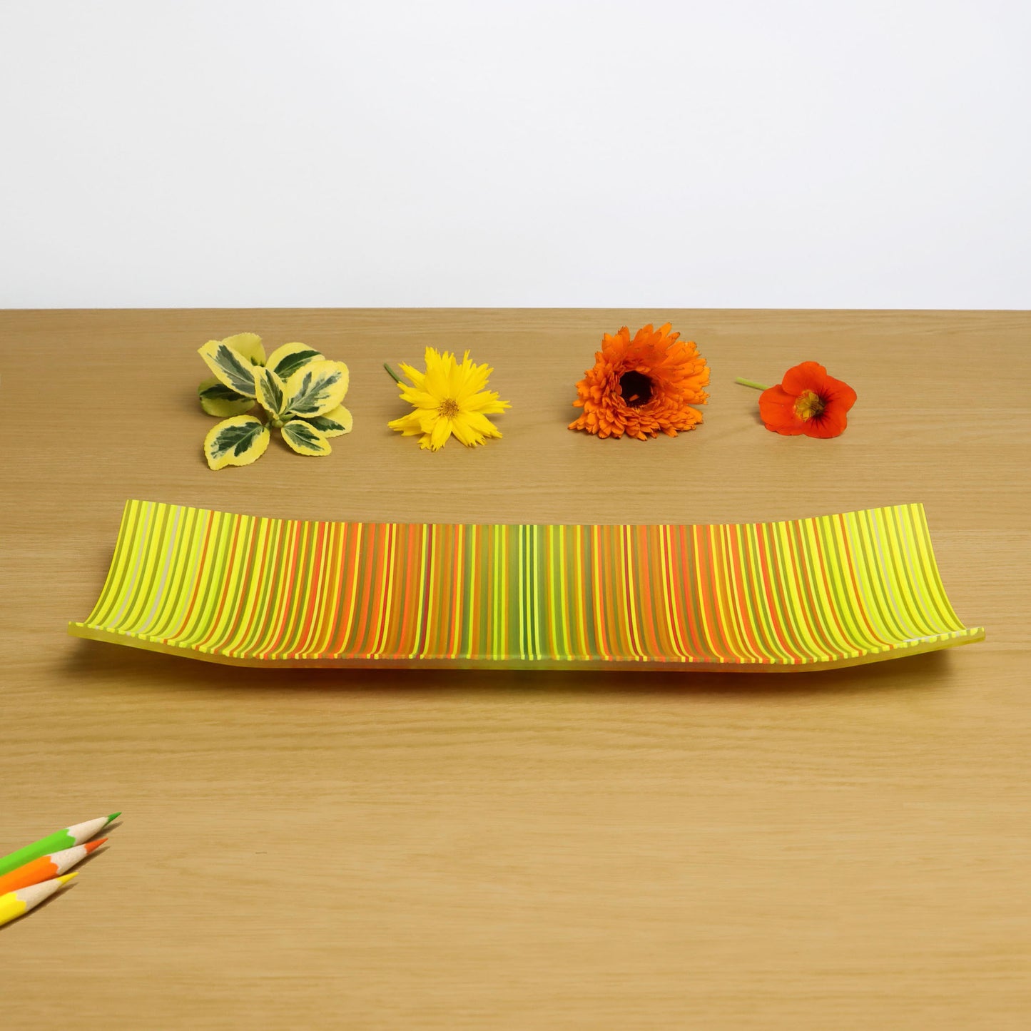 A ColourWave Glass fused glass plate displaying a radiant spectrum of colours that transition from yellow to orange to green with highlights of red amongst the sequence of coloured strands of glass. The plate’s elongated rectangular form is complemented by corners that curve gracefully upwards. It sits on a wooden surface, accentuated by flowers and leaves, all harmonising with the plate’s colour scheme. A set of coloured pencils in the foreground underscores the artistic allure of this decorative piece.