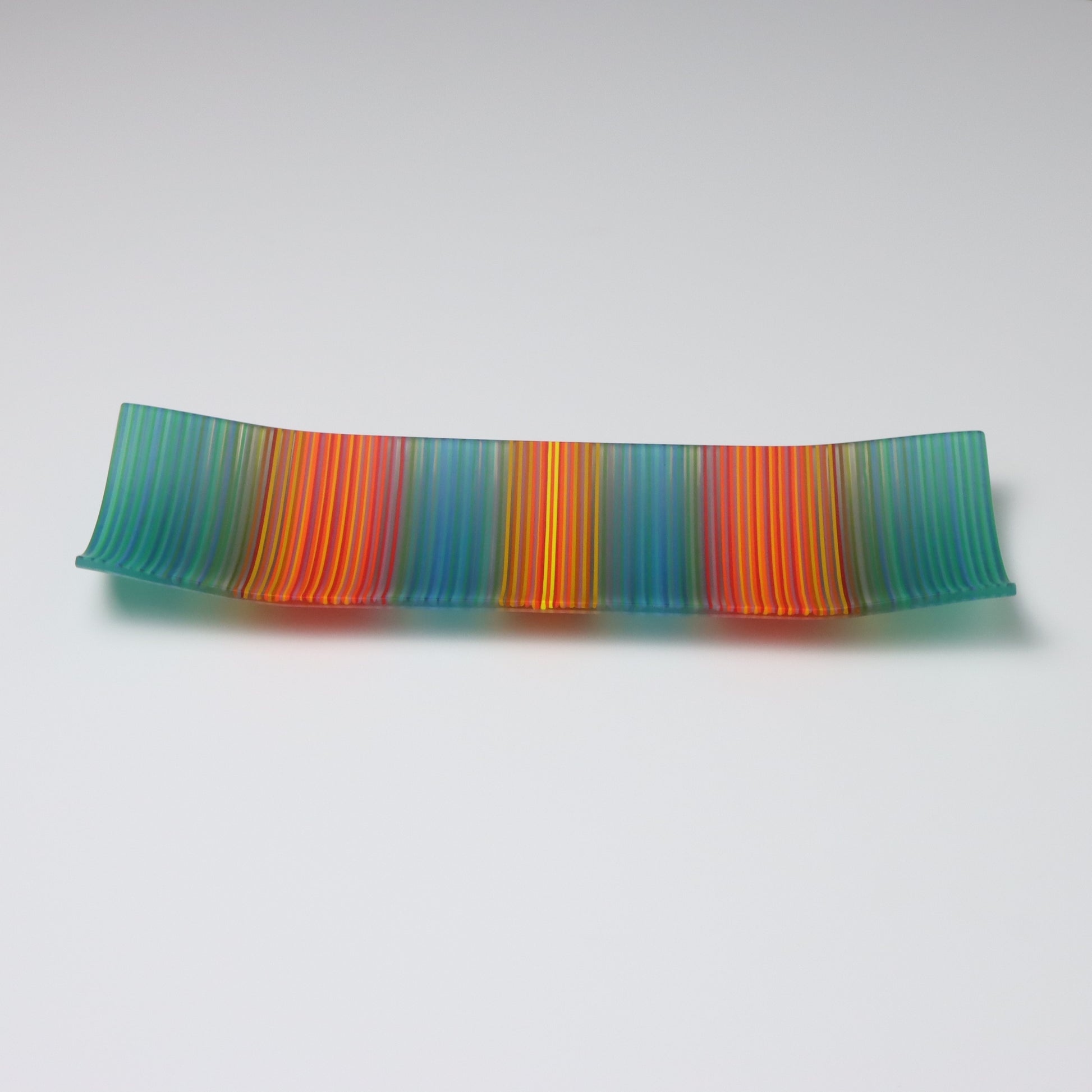 Displayed against a white backdrop is a rectangular fused glass plate from ColourWave Glass. This handcrafted piece features vertical stripes in a soothing teal green, tranquil blue, fiery red, and vibrant orange, with a radiant yellow highlight at the center. The plate’s unique design includes gentle upward curves at the corners and sides, adding a three-dimensional aspect to its visual appeal. 