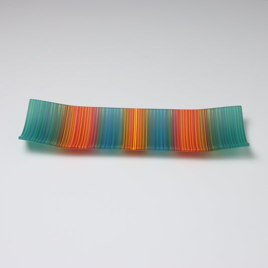 Displayed against a white backdrop is a rectangular fused glass plate from ColourWave Glass. This handcrafted piece features vertical stripes in a soothing teal green, tranquil blue, fiery red, and vibrant orange, with a radiant yellow highlight at the center. The plate’s unique design includes gentle upward curves at the corners and sides, adding a three-dimensional aspect to its visual appeal. 