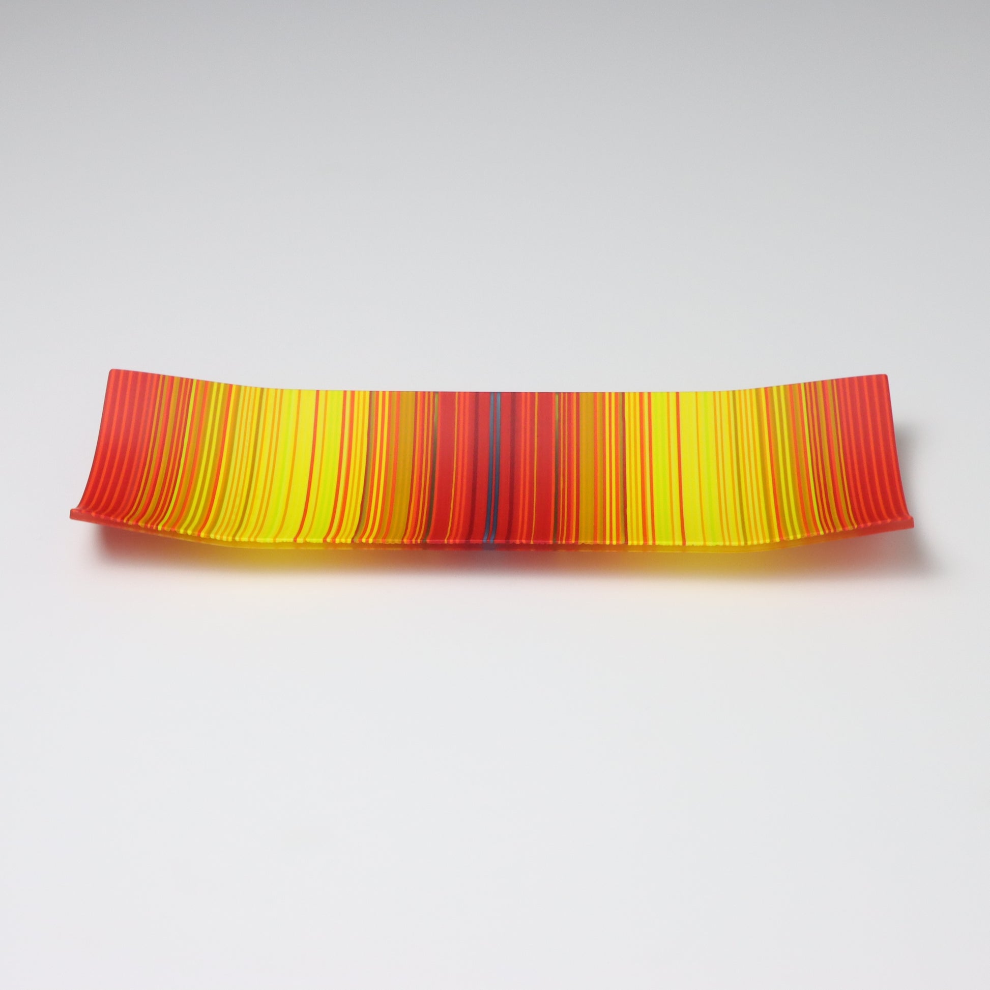 A captivating ColourWave Glass fused glass plate, showcasing a vibrant colour gradient that transitions between bright red to bright yellow. The plate’s rectangular shape is softened by the corners that curve gently upwards, adding an elegant touch to the design. Set against a neutral grey backdrop, the plate’s rich colours and sleek form are beautifully highlighted.