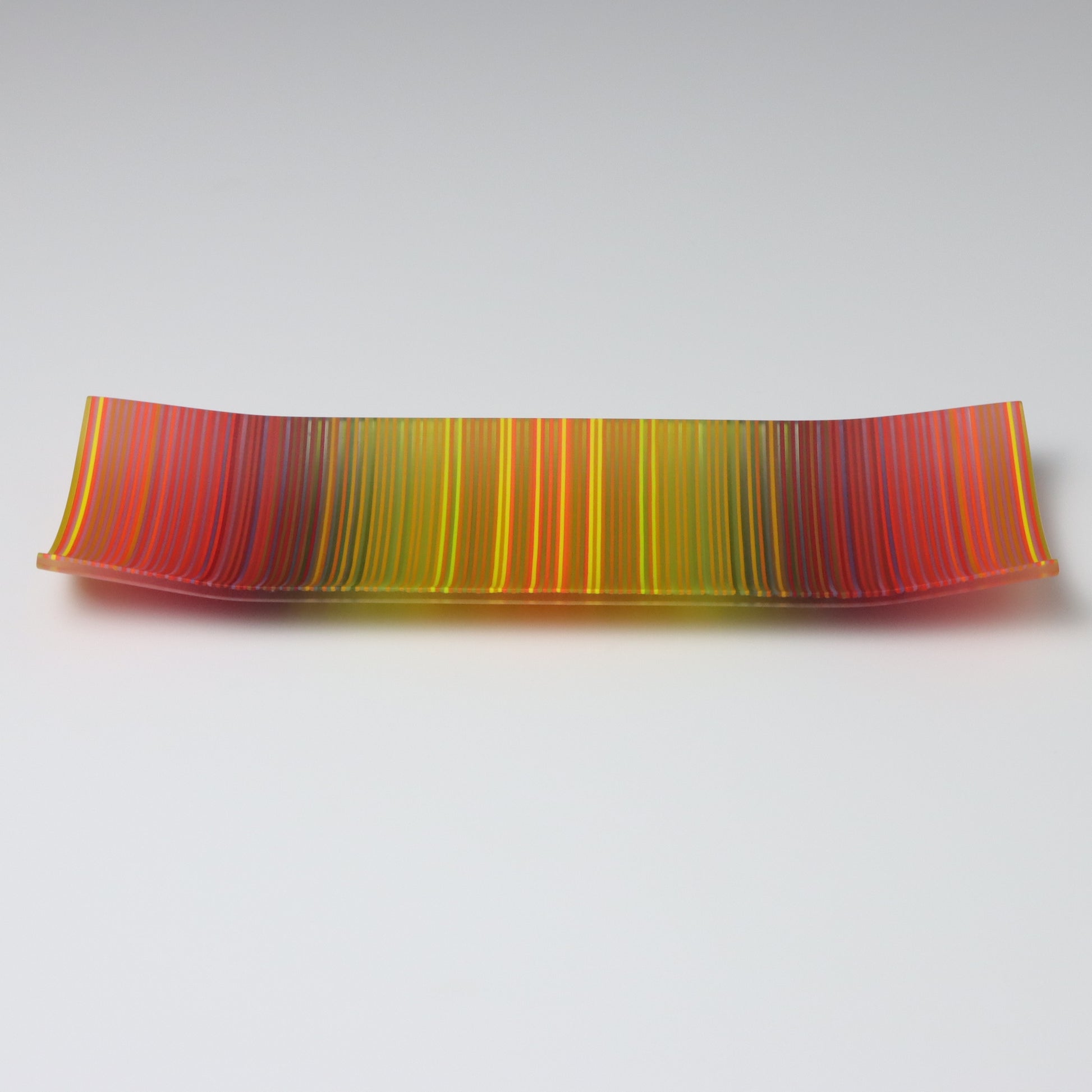 An enchanting ColourWave Glass fused glass plate, where the corners rise softly, framing a rich mosaic of autumn colours. Stripes of deep red, purple, green, and yellow, with a myriad of additional shades revealed upon close examination, create a visual celebration of autumn’s splendour.