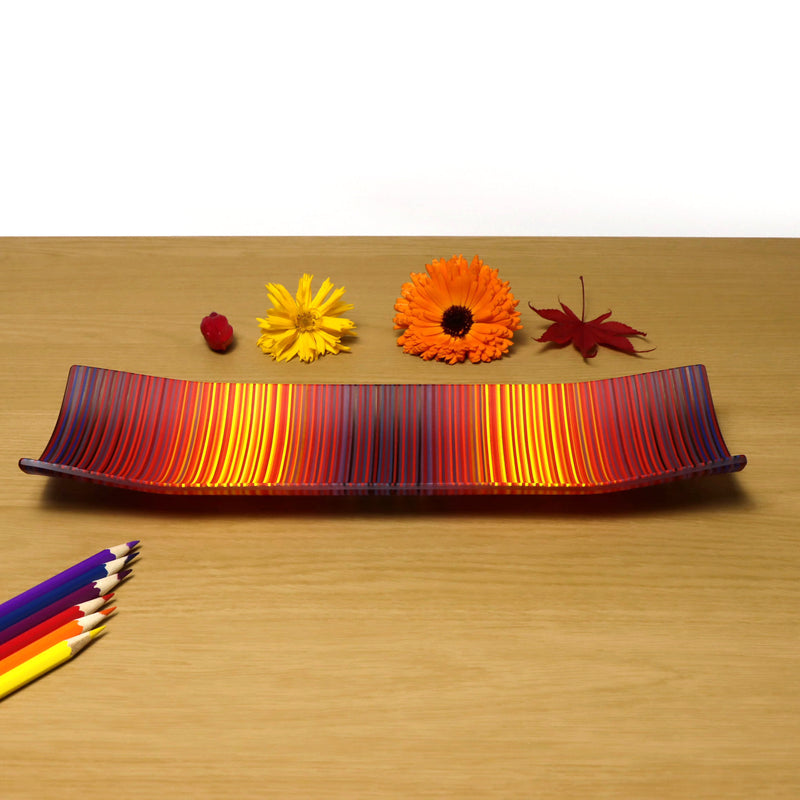 A rectangular ColourWave Glass fused glass plate, with a dynamic gradient of warm colours transitioning from deep reds to warm sunny yellows. The plate’s longer sides elegantly curve upwards at the corners. The striped pattern across the surface gives the impression of a vivid sunset, making it a striking centrepiece for that special space in your home.