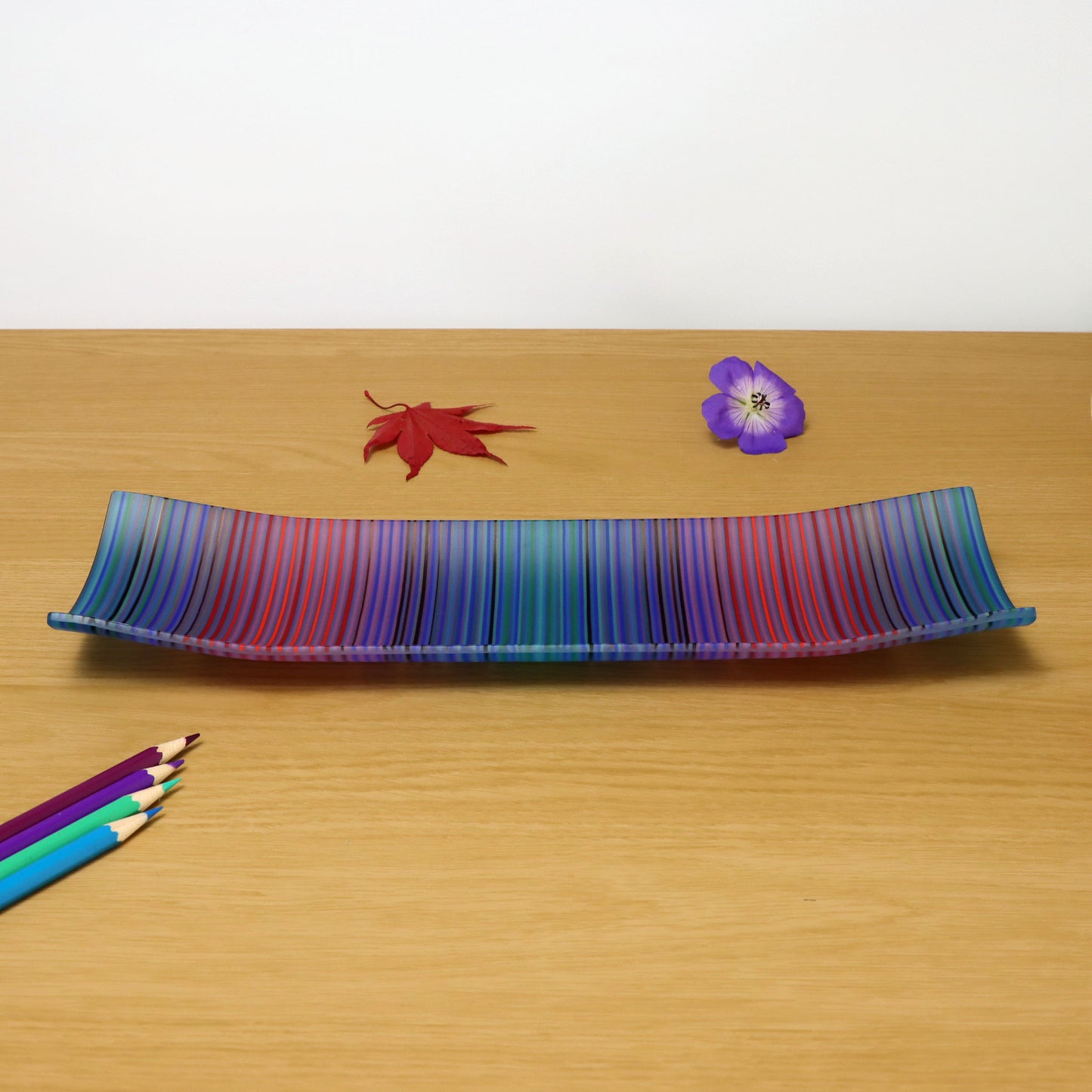A ColourWave Glass fused glass plate with vibrant vertical stripes in a spectrum of colours ranging from red, blue, green to purple. The rectangular plate features corners that gently curve upwards, giving it a sophisticated and contemporary shape. The plate is displayed on a wooden surface, accompanied by a red maple leaf, a purple flower, and a set of coloured pencils to the bottom left corner, enhancing its artistic allure.