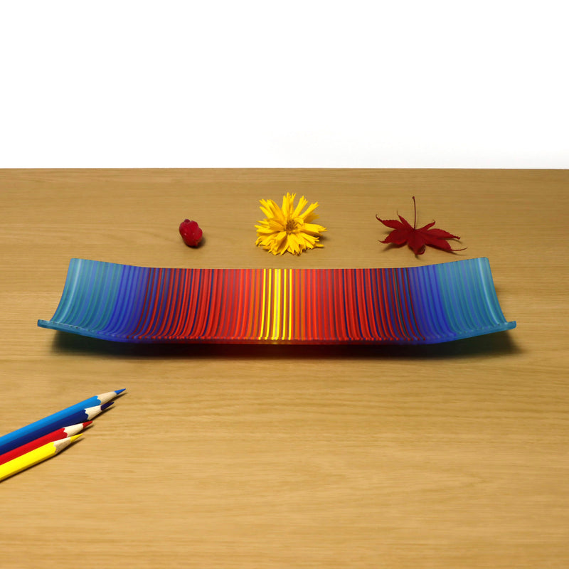 A ColourWave Glass fused glass plate featuring a vibrant spectrum of colours, with the edges curving upwards at the corners. The plate is placed on a wooden surface and is accompanied by a red berry, a yellow flower, and a red maple leaf to its right, as well as three coloured pencils to its left, adding contrast and interest to the composition.
