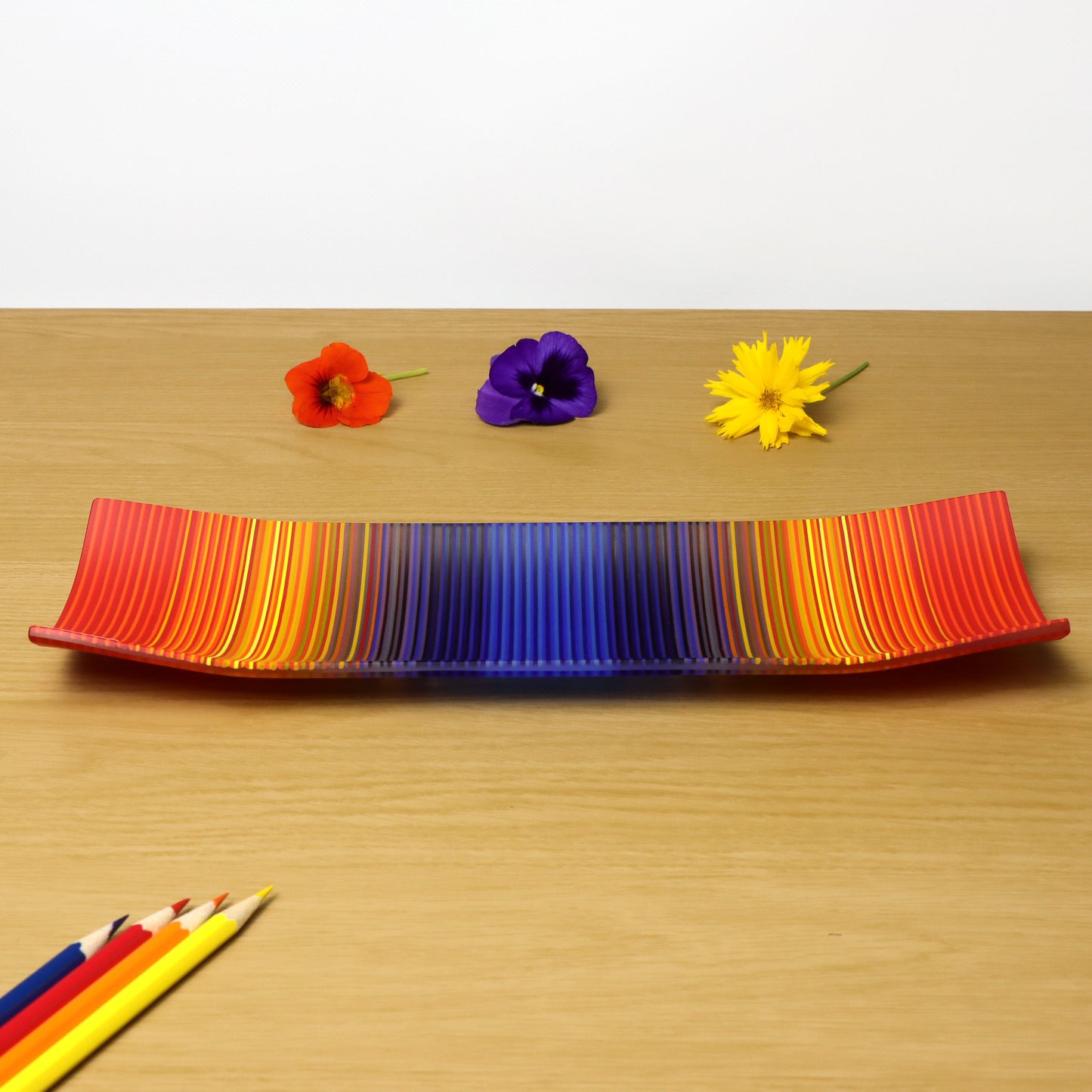 A ColourWave Glass fused glass plate, crafted in the UK, with a vibrant colour gradient. The rectangular plate features upturned corners and presents a bright blue centre that transitions into purple, yellow, and red waves of colour. The plate is displayed on a wooden surface, accompanied by a red poppy flower to the left, a purple pansy in the middle, and a yellow daisy to the right. Below the display are a set of complementary coloured pencils.