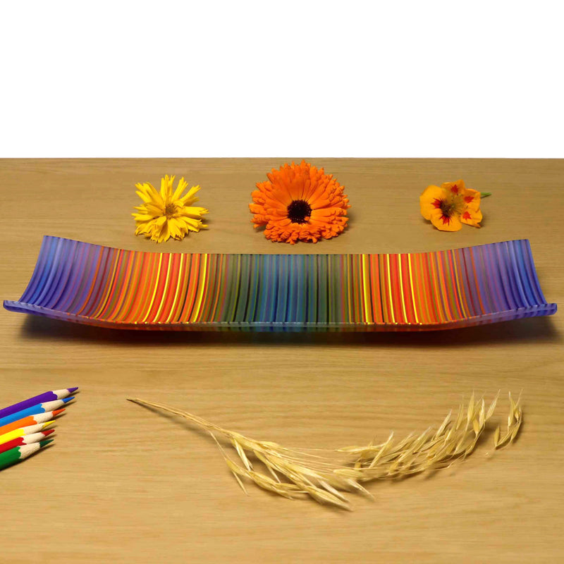 A ColourWave Glass fused glass plate, featuring a spectrum of colours including blue, orange and teal green in vertical stripes, with many more colours seen up  close. The plate has corners that elegantly curve upwards and is placed on a wooden surface, highlighted by colourful flowers and wheat stalks in the foreground, and coloured pencils placed below.