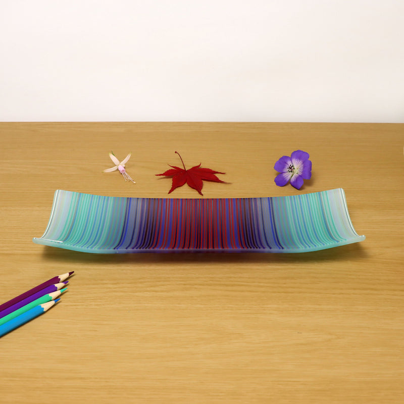 A vibrant ColourWave Glass fused glass plate, adorned with a spectrum of blue, green, purple, and red stripes. The rectangular plate subtly curves at the corners, creating a refined raised edge. It is displayed on a wooden surface, accentuated by a red maple leaf and a purple flower to its right, with an assortment of coloured pencils in the foreground.