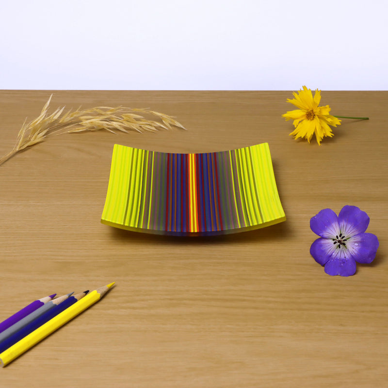 A ColourWave fused glass plate, resting on a wooden surface, displays a captivating vibrant gradation from yellow to purple, with many other colours making up the sequence. Adjacent to the plate, a yellow flower and a single stalk of wheat add a touch of nature to the scene. In the foreground, coloured pencils in matching shades of yellow purple and blue, lie diagonally pointing towards the plate, with a striking purple flower adjacent. 