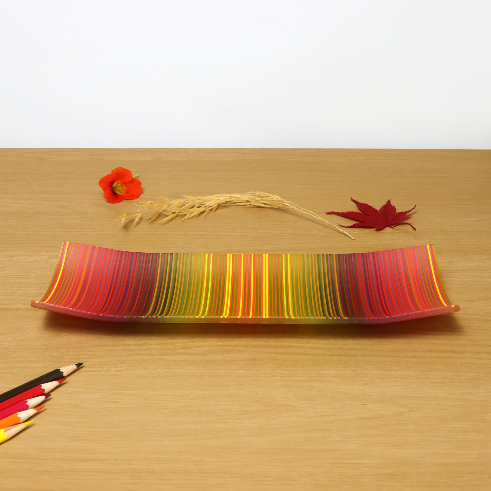 A vibrant ColourWave Glass fused glass plate with a dynamic spectrum of vertical stripes ranging from red to green. The plate’s corners elegantly curve upwards, adding a touch of sophistication. It is placed on a wooden surface, flanked by a single poppy flower to the left and a red maple leaf to the right, with a bundle of wheat stalks and four coloured pencils lying in front of it in harmonising colours.